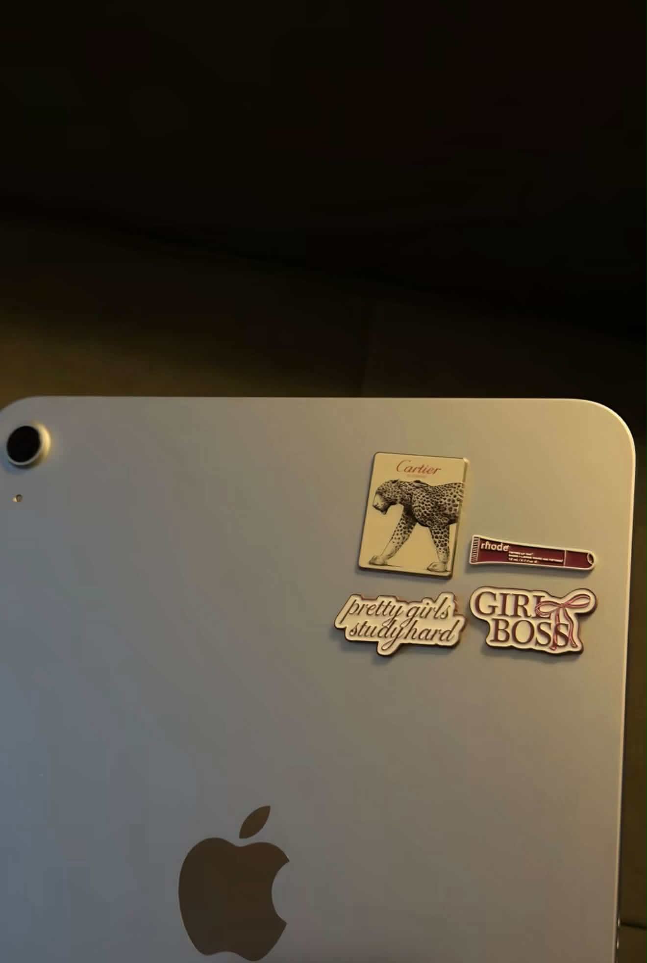 Four metal stickers clustered on the back of a silver laptop, including a leopard card, a lip gloss and two lettering stickers